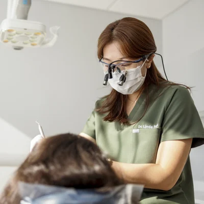 Dentist in green scrubs, mask, and magnifying glasses, focused on treating a patient under bright dental lights. Calm, professional atmosphere.